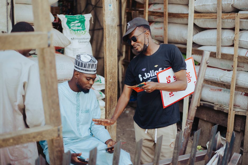 Two men interacting in an outdoor market, surrounded by sacks of goods.